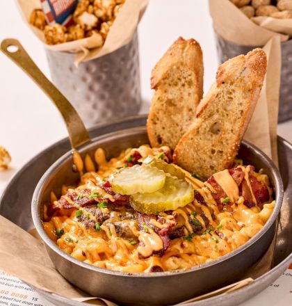 A skillet dish topped with pickles and toast, surrounded by popcorn and peanuts in metal containers on a table with a menu.