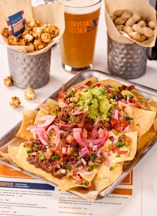 Nachos topped with beef, guacamole, and veggies, alongside popcorn, peanuts, and a beer on a table.