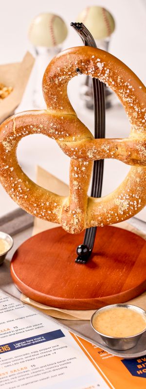 A large pretzel on a stand is served with dips, accompanied by a drink, popcorn, and a menu on a table.