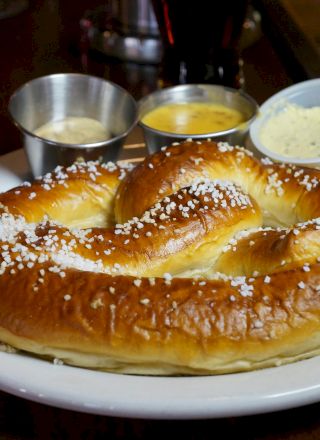 A pretzel sprinkled with salt is on a plate, accompanied by three small cups of dipping sauces.