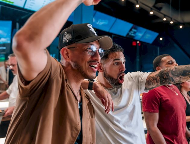 A group of people excitedly watches something, cheering with enthusiasm in a lively indoor setting.