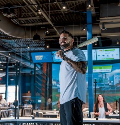 A person is indoors holding a baseball bat, with digital screens and tables around, suggesting a sports bar or entertainment venue.