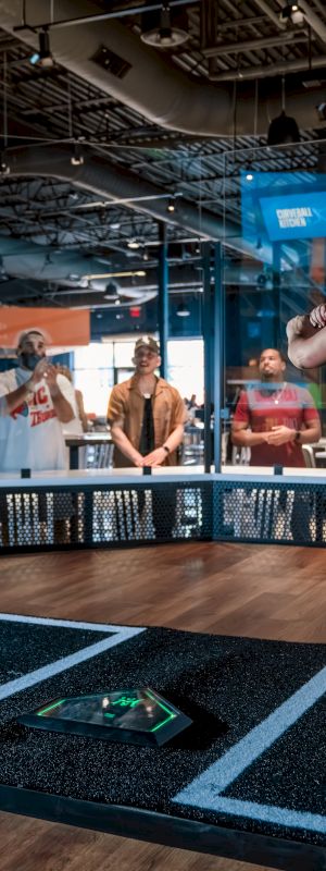 A person is playing in a virtual sports setup, with others watching in the background of a modern indoor facility.