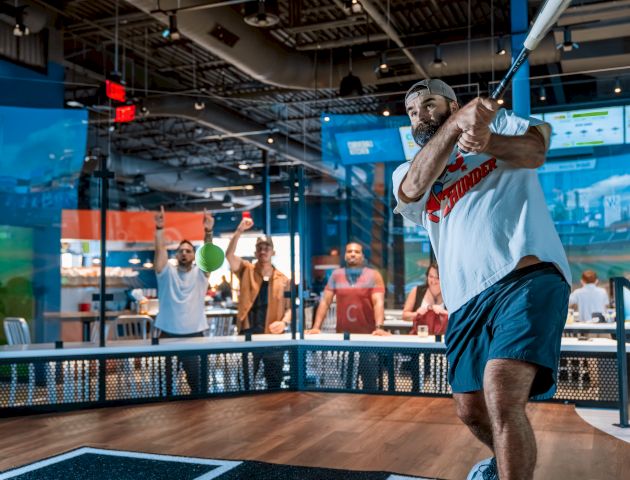 A man in a sports setting is swinging a baseball bat indoors, with a group of people watching in the background.