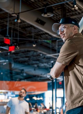 A person in a brown shirt and cap is holding a bat indoors, appearing ready to swing, as another person watches in the background.