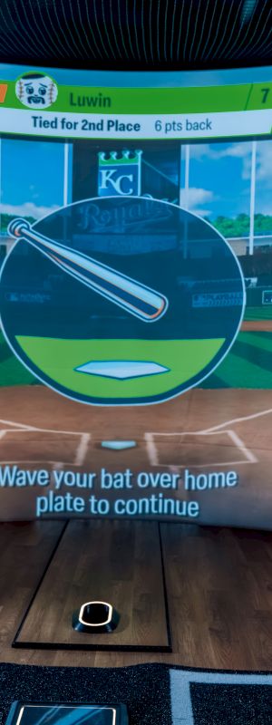 A person is playing a virtual baseball game, holding a bat in a simulator setup with a stadium displayed on the screen.