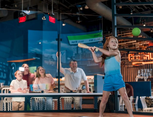 A young girl swings a baseball bat at a moving ball indoors, while a group of people seated nearby watch her play.