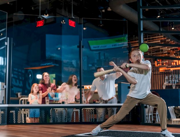 A person is swinging a bat at a green ball indoors, with onlookers behind a glass barrier cheering.