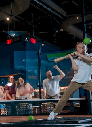 A person is playing a virtual reality game with a green bat in an arcade, while others watch excitedly in the background.