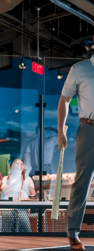 A person is playing indoor cricket, holding a bat, and looking upwards. Others watch from behind a glass panel in a lively venue.