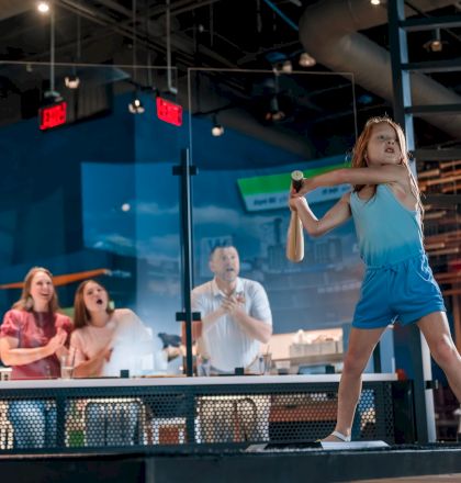 People are indoors, enthusiastically watching a girl hit a ball with a bat in a recreational setting.