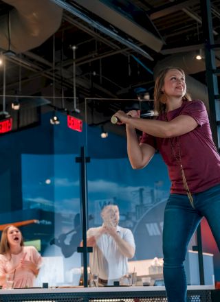A woman is playing a batting cage game indoors while onlookers watch from the side, with a lively atmosphere around.