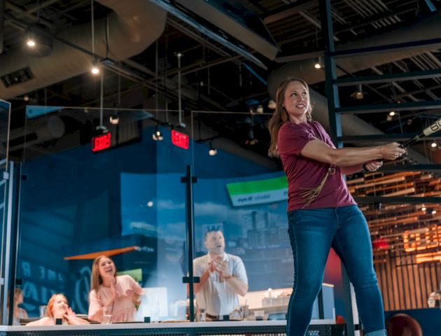 A woman poses as if swinging a bat in an indoor, modern setting, with people observing and an industrial ceiling above.