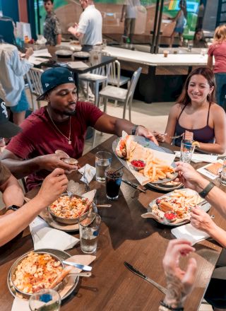 A group of people are gathered around a table sharing a meal in a casual restaurant setting, with plates of food and drinks visible.