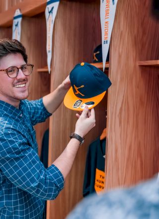 A person in a plaid shirt holds a navy and yellow cap, smiling at another person in a room with wooden cubbies and hanging lanyards.