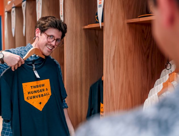 A person smiling and holding a shirt with "THROW HIGH HEAT & A CURVEBALL" printed on it in a clothing store.