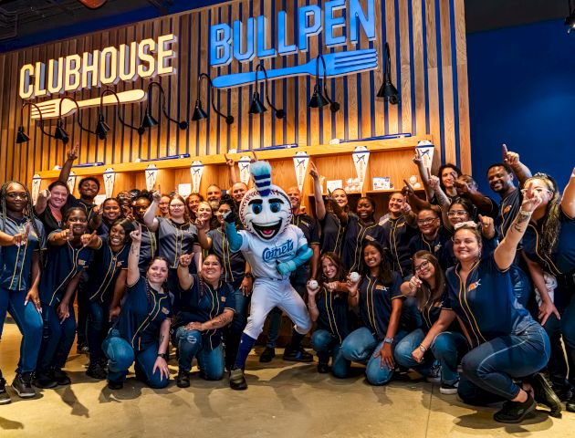 A group of people posing with a mascot in front of a "Clubhouse Bullpen" sign, all showing excitement and unity.