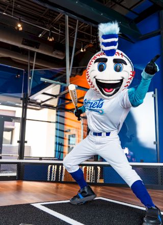 A baseball mascot in a white uniform and blue accents poses with a bat indoors, set in a sporty, vibrant environment.
