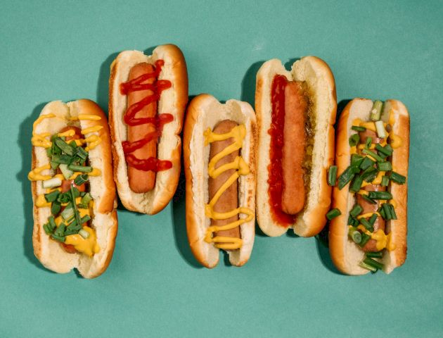 Five hot dogs with various toppings like ketchup, mustard, relish, and green onions are arranged on a green background.