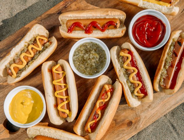 A wooden board with various hot dogs, condiments like mustard, ketchup, relish, and different toppings on a sandy surface.
