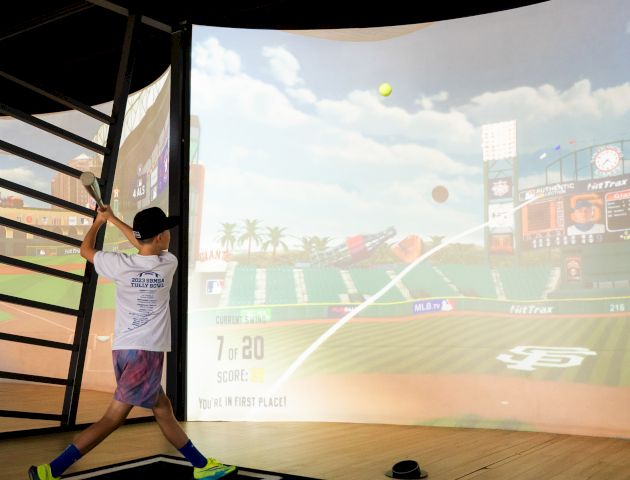 A person swings a bat in front of a virtual baseball simulation in an indoor setting.