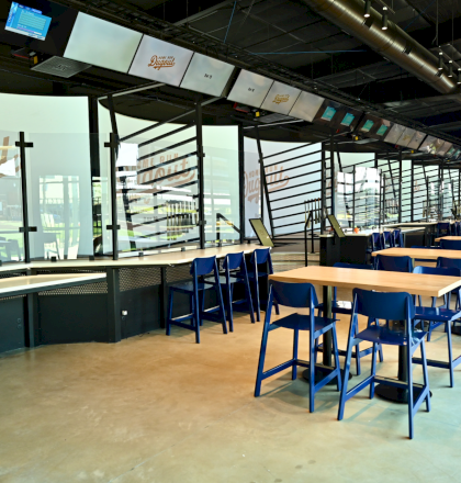 The image shows an indoor dining area with tables and chairs, featuring baseball-themed decor and digital screens overhead.