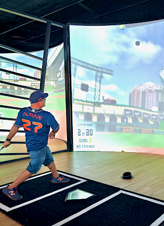 A person in a baseball uniform is playing a virtual baseball game, swinging a bat at a screen displaying a stadium.