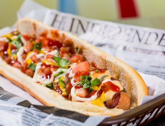 A loaded hot dog topped with tomatoes, cilantro, onions, mustard, and ketchup, in a bun, served on newspaper.