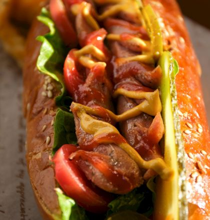 The image shows a hot dog with lettuce, sliced tomatoes, pickles, mustard, and ketchup in a bun, with waffle fries in the background.