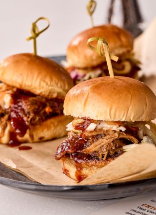 Sliders with pulled pork, coleslaw, and barbecue sauce on buns, secured with picks, served on a plate lined with paper.