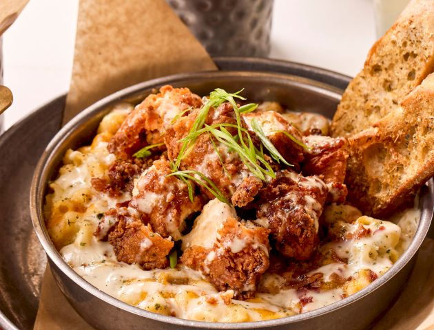 A bowl of creamy pasta topped with crispy fried chicken and herbs, served with toasted bread slices. Popcorn and a menu are in the background.