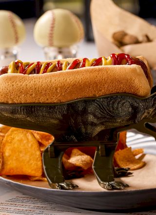 A hot dog with a toy dinosaur as a base on a plate with chips. Baseball-themed decor is in the background.