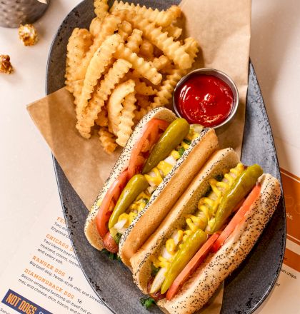 The image shows two hot dogs with pickles, tomatoes, and mustard, crinkle-cut fries with ketchup, and a menu on a plate.