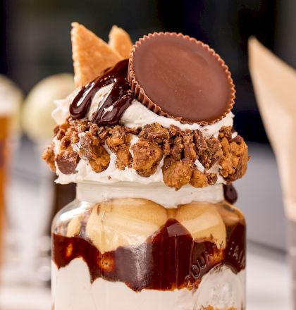 A decadent dessert in a jar featuring whipped cream, chocolate sauce, peanut butter, and a Reese's cup on top.