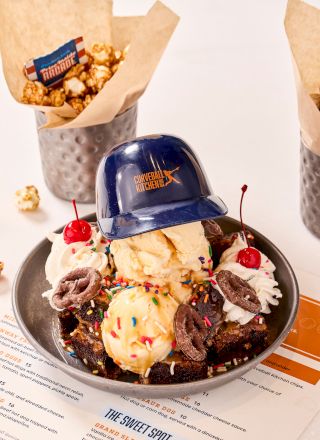 A dessert with ice cream scoops, chocolate toppings, cherries, sprinkles, and a cap. Popcorn in cups in the background.