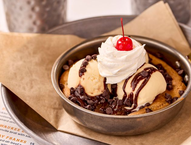 A skillet dessert with ice cream, chocolate sauce, chocolate chips, whipped cream, and a cherry on top, placed on a paper-lined tray.