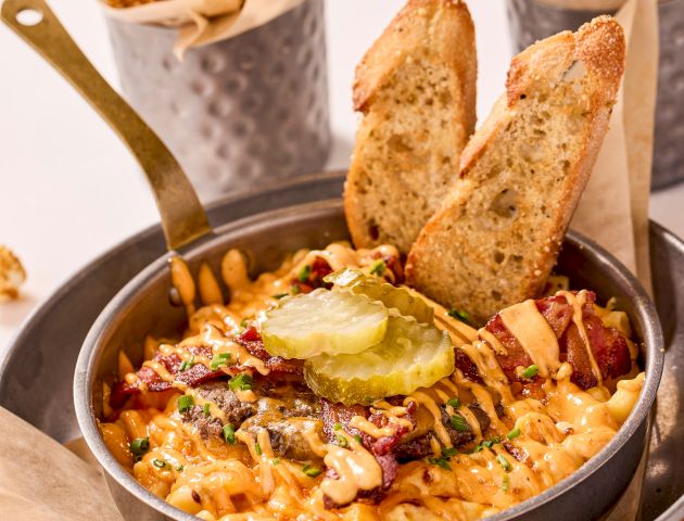 The image shows a skillet dish of loaded fries with cheese, bacon, pickles, and bread. Popcorn and peanuts in containers are in the background.