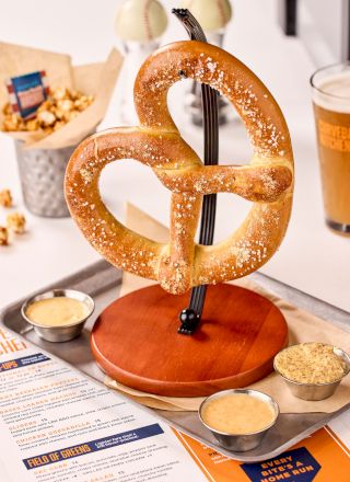 A large salted pretzel on a stand is served with dipping sauces. Nearby are popcorn, a beer glass, and a menu on the table.