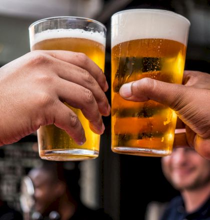 Two people are clinking glasses of beer in a celebratory gesture, with a blurred background that suggests a social setting.