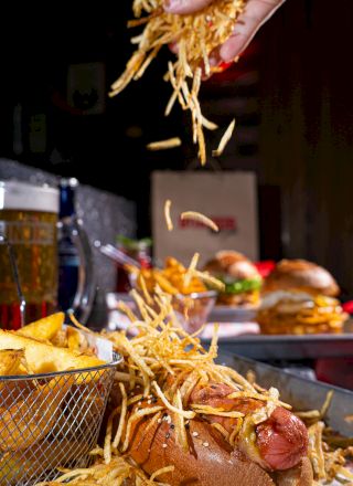 A hand sprinkles crispy fries over a loaded hot dog. Burgers and drinks are in the background, creating a classic fast-food scene.
