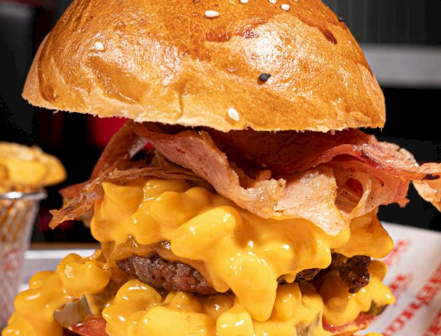 A burger topped with bacon and a generous serving of macaroni and cheese, served on a bun with sesame seeds on top.