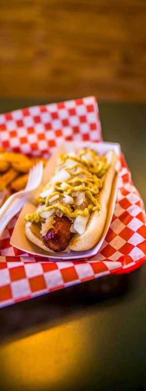A hot dog with toppings and a side of fried snacks on a red checkered paper-lined tray with a fork and knife.