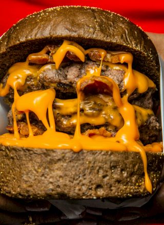 A person holds a large cheeseburger dripping with orange cheese in both hands, wrapped in paper against a red background.