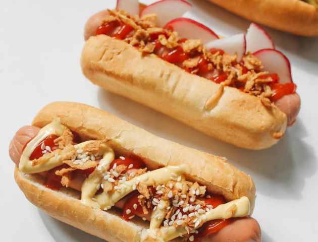 Three hot dogs with toppings like mustard, ketchup, radish slices, and crispy onions displayed on a light background.