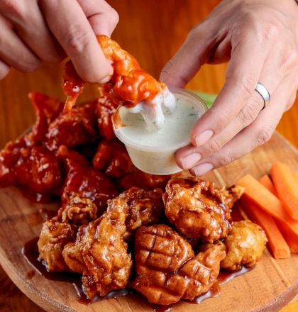 A person dips saucy chicken wings into a cup of dressing on a wooden plate alongside carrot sticks and more chicken wings.