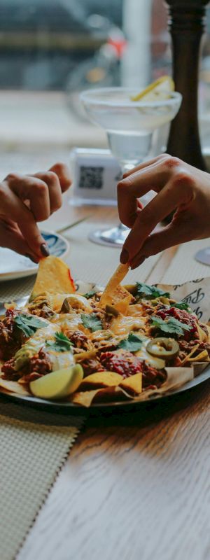 People are sharing nachos and enjoying drinks at a table, reaching over for food, in a cozy setting by the window.