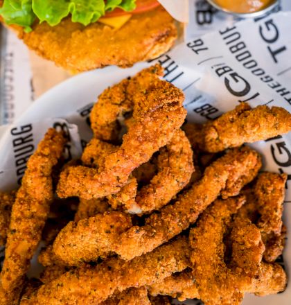 The image shows a plate of crispy chicken strips, a sandwich with lettuce, and a cup of dipping sauce on printed paper.
