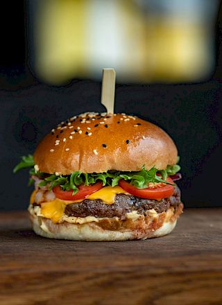 A cheeseburger with lettuce, tomato, and cheese on a sesame seed bun, secured with a wooden skewer, placed on a wooden surface.