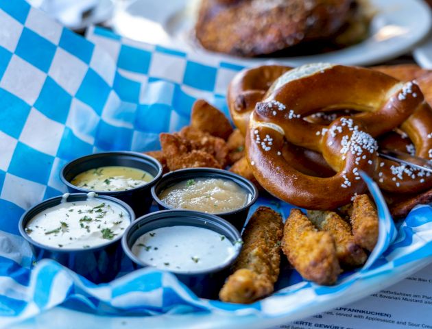 A basket with a salted pretzel, breaded sticks, dipping sauces, and fried snacks on a blue checkered paper.