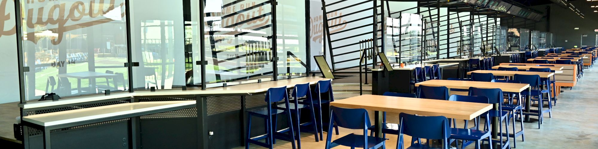 The image shows a spacious, modern dining area with tables, chairs, and a row of computer stations or kiosks along a windowed wall.
